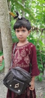 A young child is standing next to a tree, wearing a maroon traditional dress embellished with gold embroidery. They have a black bag slung over their shoulder and their hair is styled with a decorative clip. The background is a lush green natural setting.