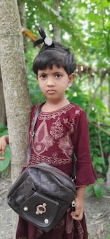 A young child is standing next to a tree, wearing a maroon traditional dress embellished with gold embroidery. They have a black bag slung over their shoulder and their hair is styled with a decorative clip. The background is a lush green natural setting.