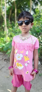 A young child stands outdoors, wearing a pink outfit with floral designs. The child is accessorized with dark sunglasses, a beaded necklace, and several pink bangles. The background consists of lush green foliage and trees.