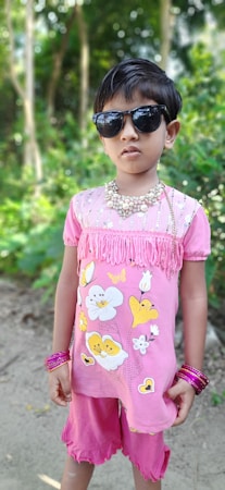 A young child stands outdoors, wearing a pink outfit with floral designs. The child is accessorized with dark sunglasses, a beaded necklace, and several pink bangles. The background consists of lush green foliage and trees.