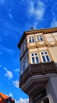 A traditional half-timbered house with ornate woodwork and white-framed windows, set against a vibrant blue sky with scattered white clouds. The triangular roof is topped with a small spire.