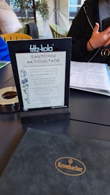 A table in a cafe or restaurant with a menu holder displaying a sign for 'Santorini Aktionstage'. The sign includes days of the week with promotional details and illustrations of drinks. There is a black folder with 'Krombacher' embossed on it and a person holding a phone nearby, along with various furniture and decor elements in the background.