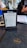A table in a cafe or restaurant with a menu holder displaying a sign for 'Santorini Aktionstage'. The sign includes days of the week with promotional details and illustrations of drinks. There is a black folder with 'Krombacher' embossed on it and a person holding a phone nearby, along with various furniture and decor elements in the background.
