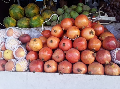 A variety of fresh fruits are displayed at a market stall. The foreground features a large pile of ripe pomegranates, with their deep red skin and distinctive crowns. To the left, apples are wrapped in protective netting, among other fruits. Behind these, green papayas and other fruits are stacked. The arrangement suggests a vibrant and colorful market setting.