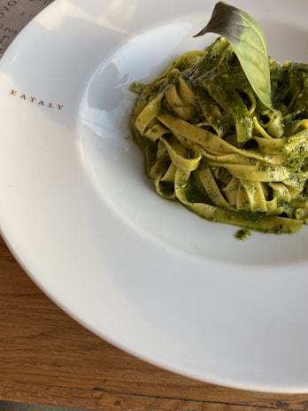 A neatly plated serving of pasta with green pesto sauce sits on a white dish marked with the word 'EATALY.' A single basil leaf is placed on top of the pasta, which rests on a wooden table surface.