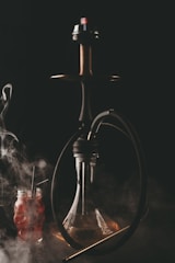 A hookah with a dark hose is depicted, emitting smoke against a dark background. Below the hookah is a clear glass base, and to the side, there is a red beverage in a jar with two straws. The scene is enveloped in swirling smoke.