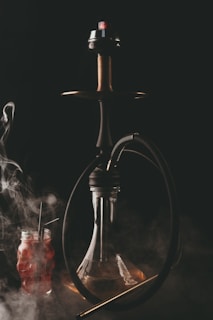 A hookah with a dark hose is depicted, emitting smoke against a dark background. Below the hookah is a clear glass base, and to the side, there is a red beverage in a jar with two straws. The scene is enveloped in swirling smoke.