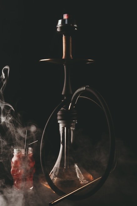 A hookah with a dark hose is depicted, emitting smoke against a dark background. Below the hookah is a clear glass base, and to the side, there is a red beverage in a jar with two straws. The scene is enveloped in swirling smoke.