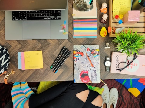 A flat lay of a colorful workspace featuring a laptop, various stationery items, a vibrant notebook with a pop art design, pens, post-it notes, and decorative items like small figurines and a potted plant. The background has a patterned table surface, and part of a seated person&rsquo;s colorful striped socks and ripped jeans are visible at the bottom. Accessories like a watch and glasses add to the organized clutter on the desk.