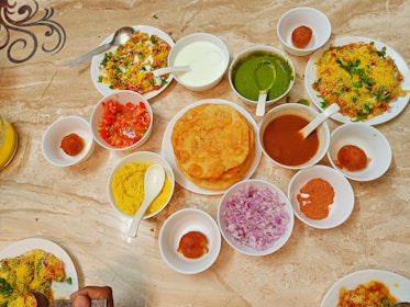 A table is filled with various Indian dishes served in small bowls and plates. The dishes include deep-fried puris, green chutney, onion slices, tomato pieces, a sweet syrupy ball dessert, and a variety of vibrant sauces. The table surface is marble, and the arrangement suggests a traditional Indian meal setup.