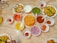 A table is filled with various Indian dishes served in small bowls and plates. The dishes include deep-fried puris, green chutney, onion slices, tomato pieces, a sweet syrupy ball dessert, and a variety of vibrant sauces. The table surface is marble, and the arrangement suggests a traditional Indian meal setup.