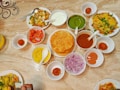 A table is filled with various Indian dishes served in small bowls and plates. The dishes include deep-fried puris, green chutney, onion slices, tomato pieces, a sweet syrupy ball dessert, and a variety of vibrant sauces. The table surface is marble, and the arrangement suggests a traditional Indian meal setup.