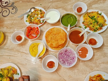 A table is filled with various Indian dishes served in small bowls and plates. The dishes include deep-fried puris, green chutney, onion slices, tomato pieces, a sweet syrupy ball dessert, and a variety of vibrant sauces. The table surface is marble, and the arrangement suggests a traditional Indian meal setup.