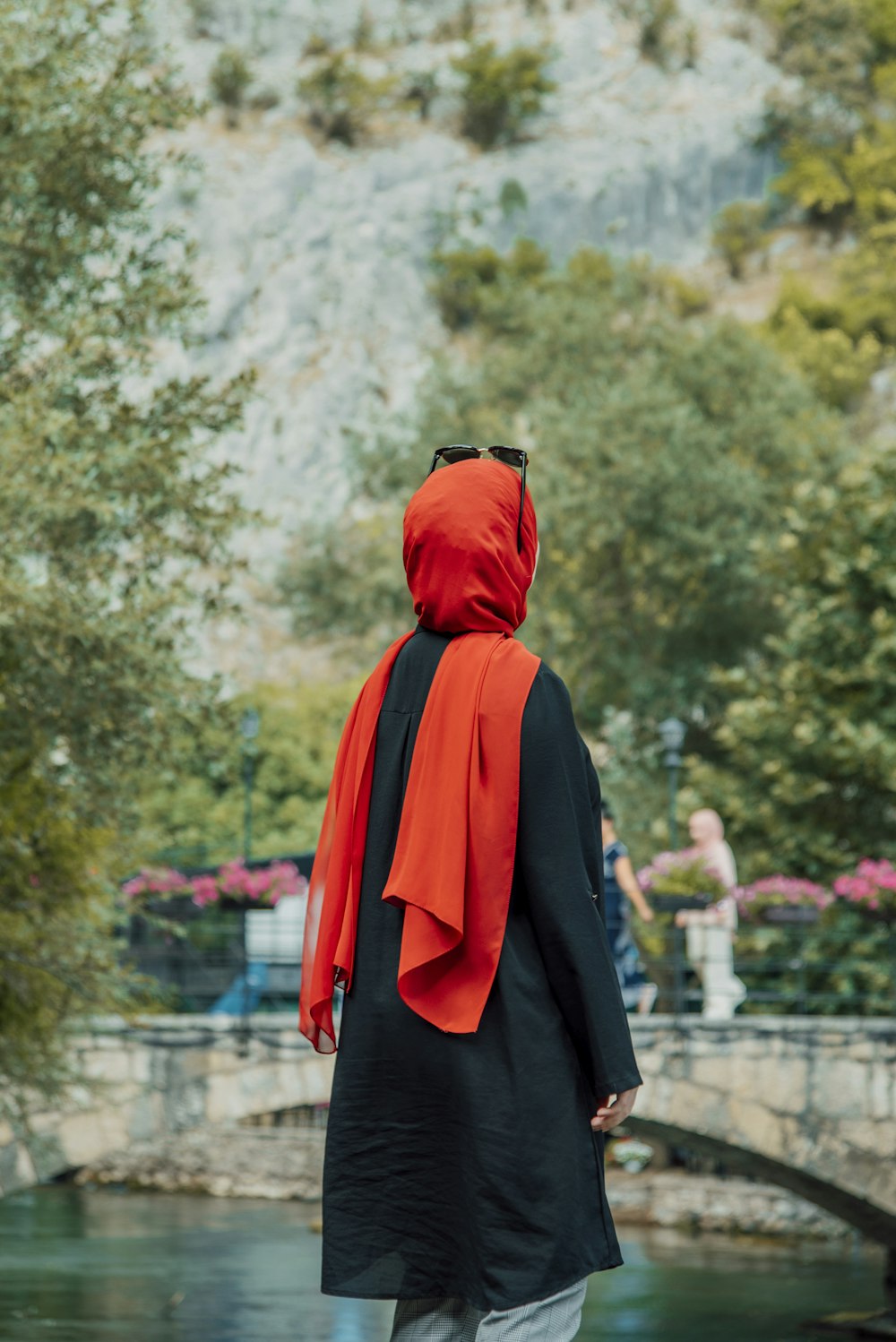 a person wearing a black robe and red head covering standing in front of a pond with trees and