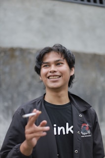 A young person with dark hair smiles while holding a cigarette. They are wearing a black jacket with a design on it and a black T-shirt with white lettering. The background appears to be a textured gray wall.