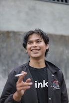 A young person with dark hair smiles while holding a cigarette. They are wearing a black jacket with a design on it and a black T-shirt with white lettering. The background appears to be a textured gray wall.
