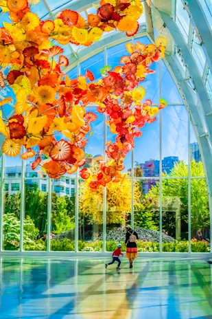 A vibrant display of colorful glass sculptures hanging from the ceiling of a spacious, sunlit conservatory. Large windows showcase a lush garden outside, with tall trees and shrubs. Two people, a child and an adult, are seen walking across the shiny floor, casting long shadows.