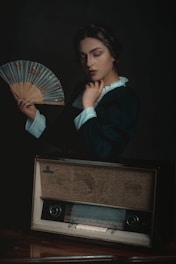 A person with a thoughtful expression holds a decorative fan. They are dressed in period-style clothing with a high-collared shirt. In front of them is a vintage radio, which adds an element of nostalgia.