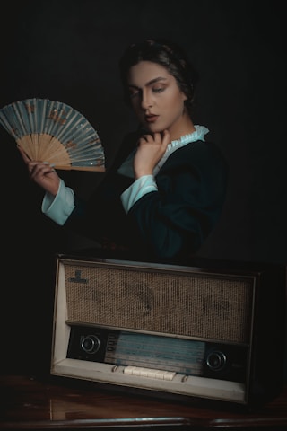 A person with a thoughtful expression holds a decorative fan. They are dressed in period-style clothing with a high-collared shirt. In front of them is a vintage radio, which adds an element of nostalgia.