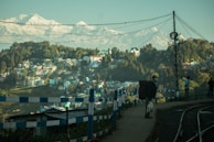A scenic view of a hillside town with numerous houses scattered across the landscape, surrounded by lush green trees. In the foreground, there is a pathway running alongside a railway track, with a few people, one carrying a large backpack and another taking a photo. The background features a majestic range of snow-capped mountains under a clear sky.