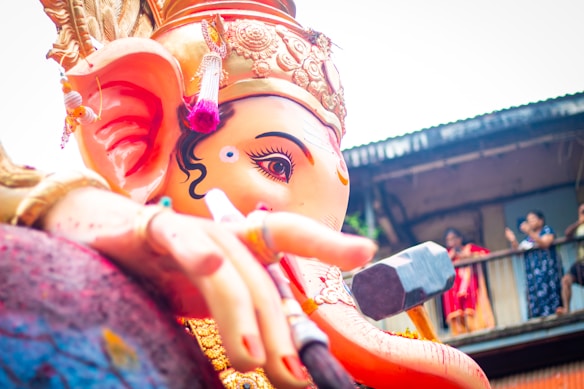 A large, colorful statue of Ganesha, an elephant-headed deity, is prominently displayed. The statue is adorned with intricate ornaments and a crown, with its hand holding a modak and an axe. There are people in the background observing the statue from the balcony of a building.