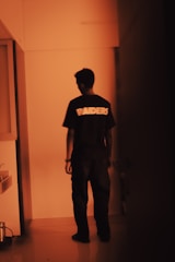 A person stands facing away in a dimly lit hallway, wearing a black shirt with the word 'RAIDERS' printed on the back. The lighting in the scene is warm and orange-tinted, casting shadows around the figure and creating a moody atmosphere.