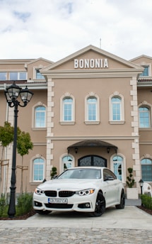 A beige building with multiple arched windows and the name 'Bononia' displayed prominently on top. In front of the building, a white BMW car with black rims is parked. The scene includes a cobblestone path, a black lamppost, and some green foliage.
