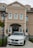 A white BMW car is parked in front of a building with beige walls and multiple windows, featuring arched designs. The building has a sign at the top with the word 'BONONIA'. There is a black lamp post on the left, and some green shrubs surrounding the area.