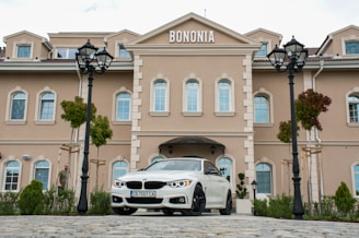 A beige, multi-story building with ornamental design and white-framed windows stands prominently. A white car with a visible license plate is parked in front of the building. There are two black street lamps and some green plants and trees surrounding the area. The word 'BONONIA' is displayed on the building facade.