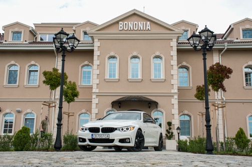 A beige, multi-story building with ornamental design and white-framed windows stands prominently. A white car with a visible license plate is parked in front of the building. There are two black street lamps and some green plants and trees surrounding the area. The word 'BONONIA' is displayed on the building facade.