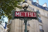 An ornate cast iron metro sign with the word 'METRO' written in white on a red background. The sign is mounted on a decorative pole with intricate detailing, topped by a spherical lamp. Behind the sign, there is a typical Parisian-style building with wrought-iron balconies, arched windows, and beige stone walls. Tree leaves are visible in the upper left corner, suggesting a pleasant, urban environment.