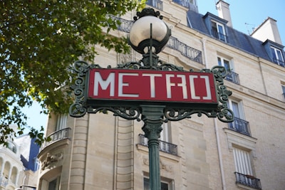 An ornate cast iron metro sign with the word 'METRO' written in white on a red background. The sign is mounted on a decorative pole with intricate detailing, topped by a spherical lamp. Behind the sign, there is a typical Parisian-style building with wrought-iron balconies, arched windows, and beige stone walls. Tree leaves are visible in the upper left corner, suggesting a pleasant, urban environment.