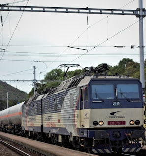 A cargo train with multiple carriages travels on a railway track. The train engine, labeled 'ZSSK Cargo,' has a distinct blue and gray color scheme. Overhead power lines are visible against a cloudy sky, with some greenery and trees in the background.