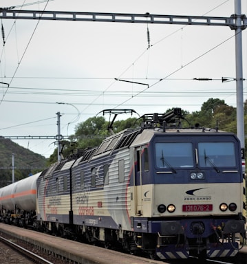 A cargo train with multiple carriages travels on a railway track. The train engine, labeled 'ZSSK Cargo,' has a distinct blue and gray color scheme. Overhead power lines are visible against a cloudy sky, with some greenery and trees in the background.