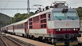 A train with a red and white locomotive is stationed on a railway track. The driver can be seen peering out of the locomotive window. Passengers are visible on the platform, appearing to board or disembark from the train. Overhead power lines and hilly terrain with trees are in the background.