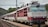A train with a red and white locomotive is stationed on a railway track. The driver can be seen peering out of the locomotive window. Passengers are visible on the platform, appearing to board or disembark from the train. Overhead power lines and hilly terrain with trees are in the background.