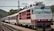 A train with a red and white locomotive is stationed on a railway track. The driver can be seen peering out of the locomotive window. Passengers are visible on the platform, appearing to board or disembark from the train. Overhead power lines and hilly terrain with trees are in the background.