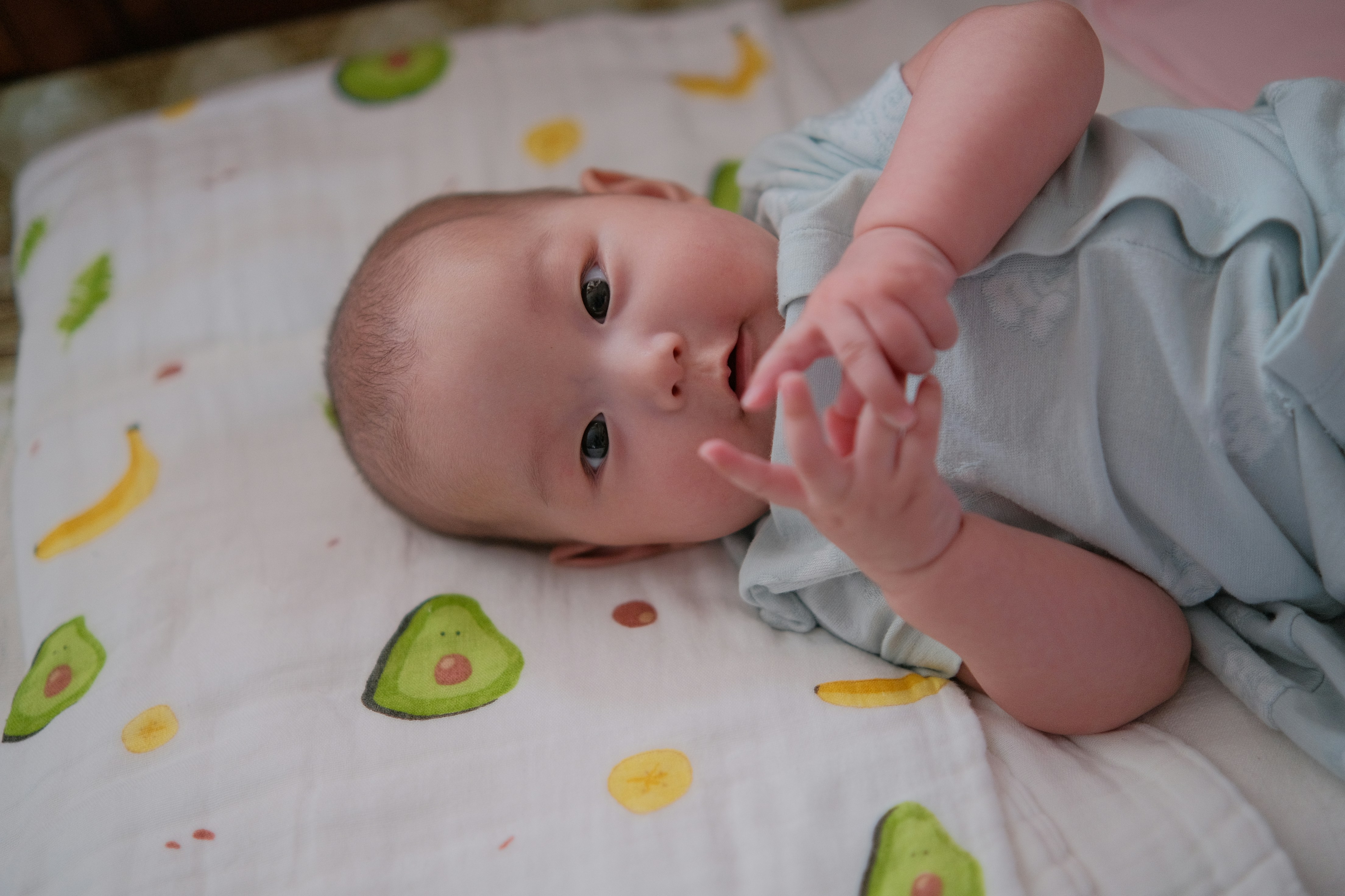 Baby lying awake on their back on a fitted sheet printed with avocados and bananas, hands near their mouth
