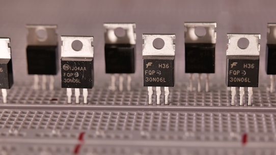 A series of identical electronic components are aligned on a breadboard. These components have a metallic top with a hole for mounting and three legs for circuit integration. The black body of the components is labeled with various alphanumeric codes.