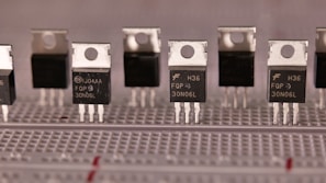 A series of identical electronic components are aligned on a breadboard. These components have a metallic top with a hole for mounting and three legs for circuit integration. The black body of the components is labeled with various alphanumeric codes.