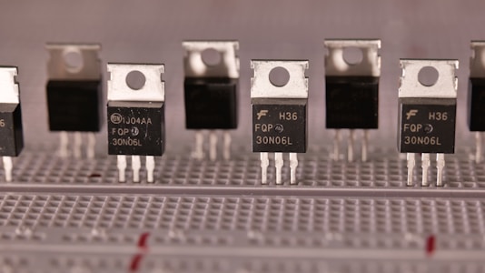 A series of identical electronic components are aligned on a breadboard. These components have a metallic top with a hole for mounting and three legs for circuit integration. The black body of the components is labeled with various alphanumeric codes.