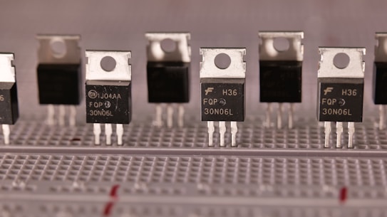 A series of identical electronic components are aligned on a breadboard. These components have a metallic top with a hole for mounting and three legs for circuit integration. The black body of the components is labeled with various alphanumeric codes.