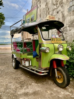 A colorful tuk-tuk taxi is parked next to an old building. The tuk-tuk is primarily green with yellow and red accents and has a black canopy roof. It is adorned with signs and a license plate in a foreign script. The background includes a textured concrete wall and some greenery.