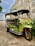 A colorful tuk-tuk taxi is parked next to an old building. The tuk-tuk is primarily green with yellow and red accents and has a black canopy roof. It is adorned with signs and a license plate in a foreign script. The background includes a textured concrete wall and some greenery.