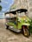 A colorful tuk-tuk taxi is parked next to an old building. The tuk-tuk is primarily green with yellow and red accents and has a black canopy roof. It is adorned with signs and a license plate in a foreign script. The background includes a textured concrete wall and some greenery.