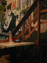 The image features the interior of a cafe or restaurant, showcasing urban art on the walls. A mix of artwork, including a painting of a skeletal figure in a military hat embracing a woman, fills the space under a wooden staircase. A framed picture of a person in red clothing is visible against the wall. A table with a clear bottle and a black tissue box is situated at the center, with green plants hanging around the stairs.