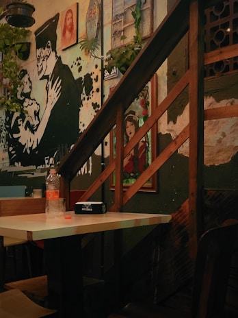 The image features the interior of a cafe or restaurant, showcasing urban art on the walls. A mix of artwork, including a painting of a skeletal figure in a military hat embracing a woman, fills the space under a wooden staircase. A framed picture of a person in red clothing is visible against the wall. A table with a clear bottle and a black tissue box is situated at the center, with green plants hanging around the stairs.