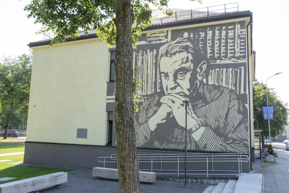 A mural painted on the side of a building features a pensive figure with folded hands and a backdrop of books. The artwork uses large, bold lines in a monochrome palette. The scene takes place in a small urban setting with a tree partially obscuring the image and a clear sky overhead.