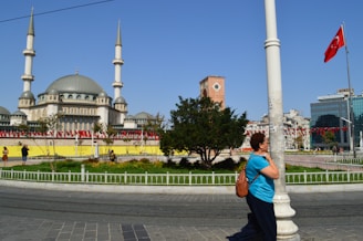 A large mosque with two tall minarets dominates the background, while a woman in a blue shirt and black pants stands next to a light pole. A red flag with a crescent and star is prominently displayed on a flagpole. There are people walking and taking photos in the plaza, which is surrounded by a circular garden with trees and flowers. In the background, there are modern buildings and a clock tower.