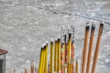 A collection of incense sticks in varying sizes and colors, with some featuring decorative patterns and characters. The sticks are arranged in a holder on a concrete surface, and a few of them are partially burned, emitting faint smoke.